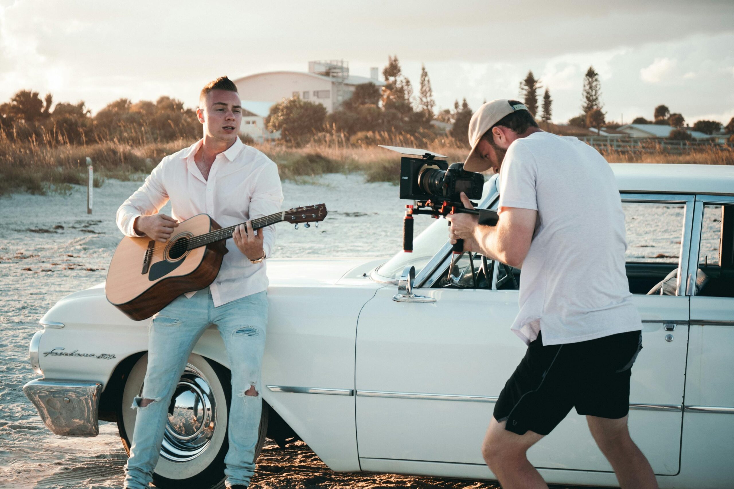 A young man plays guitar as a videographer films by a vintage white car on a sunny beach day.